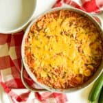 A skillet filled with a cheesy vegetable casserole, topped with melted cheese, next to a checkered red and white cloth and three whole zucchinis.