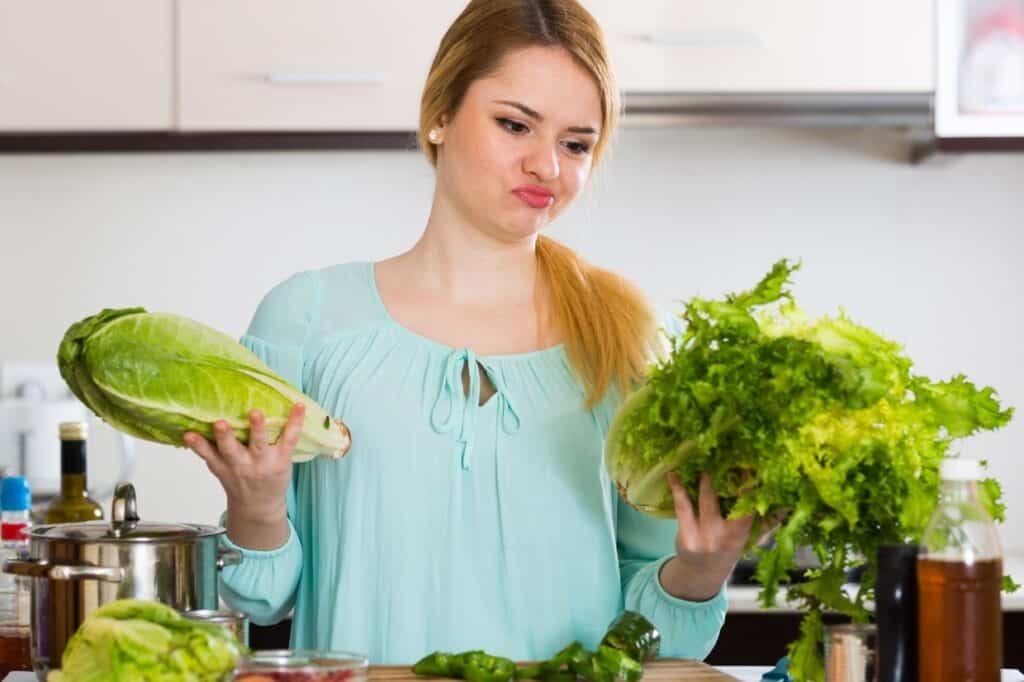 A woman in a kitchen looks at a head of lettuce and a bunch of leafy greens, appearing unsure while holding each in her hands.