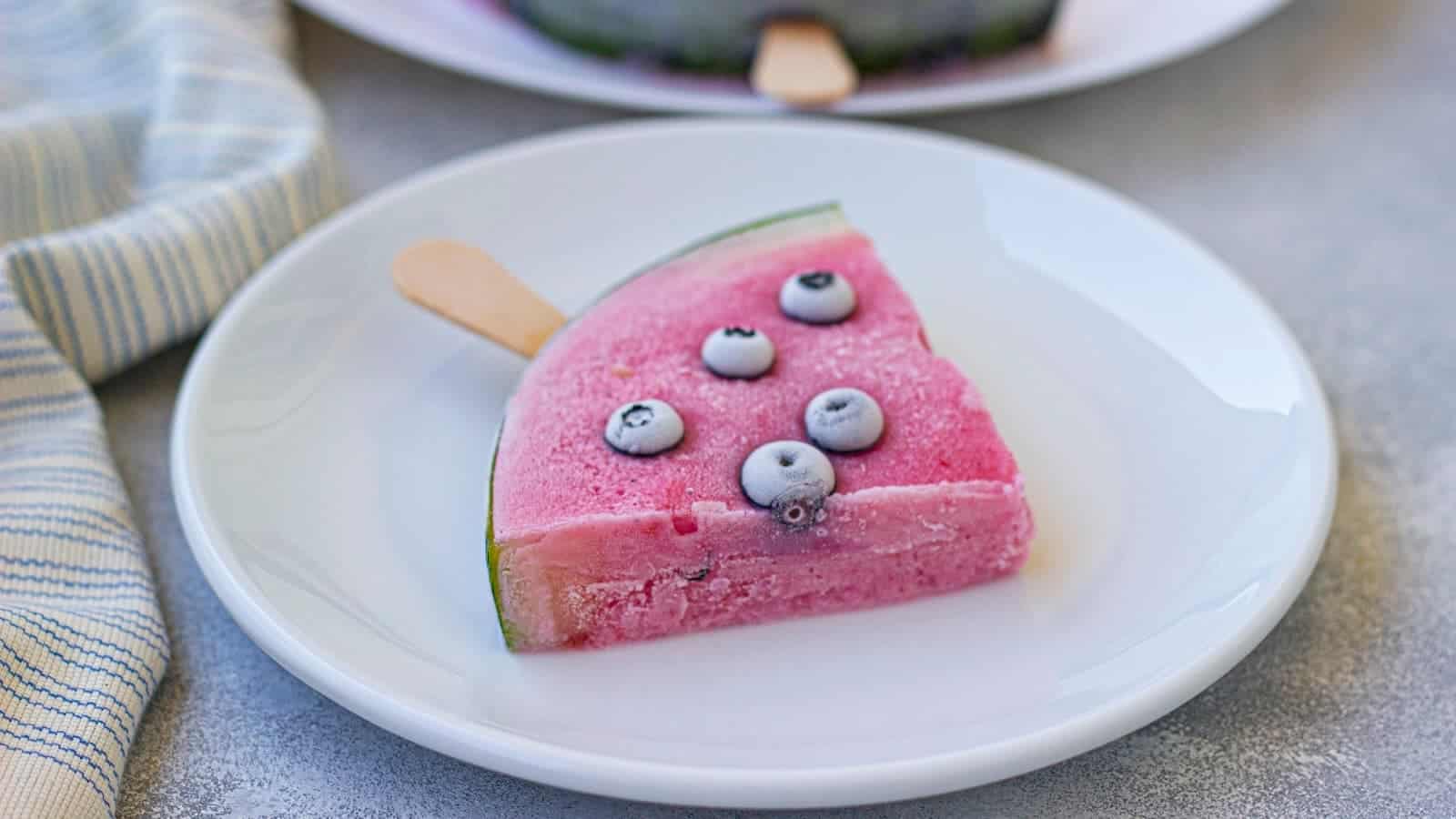 A slice of frozen watermelon wedge with blueberries, served on a white plate next to a striped napkin.