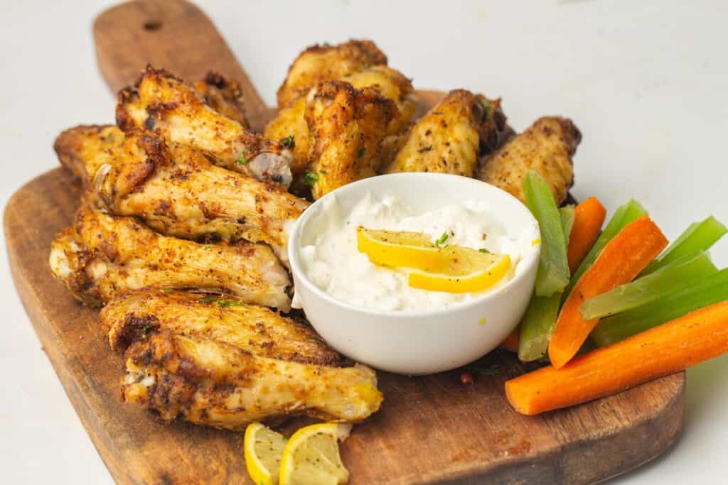 A wooden board with seasoned chicken wings, a bowl of white dipping sauce with lemon slices, and celery and carrot sticks.