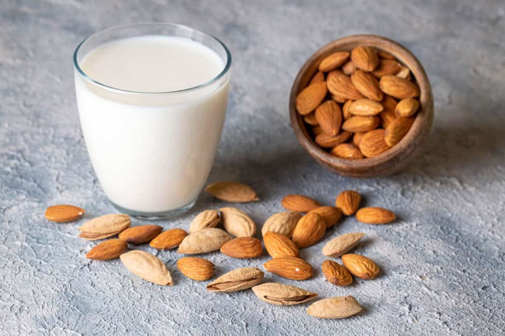 A glass of almond milk sits beside a tipped wooden bowl with whole almonds spilling out on a textured gray surface.