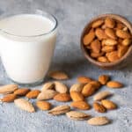 A glass of almond milk sits beside a tipped wooden bowl with whole almonds spilling out on a textured gray surface.