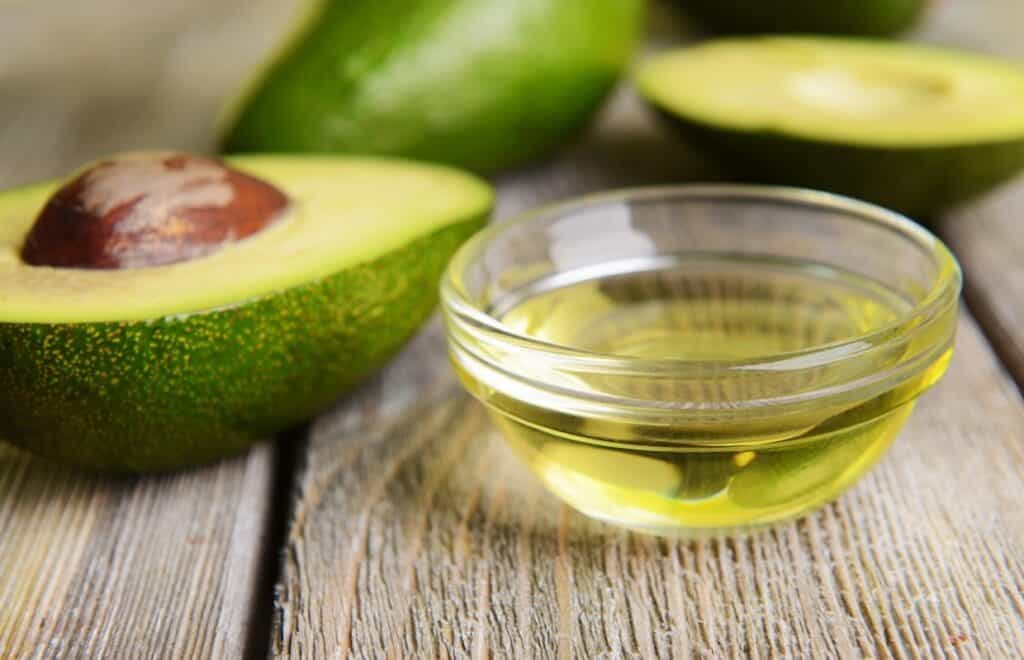 A small glass bowl of avocado oil next to halved avocados with pits, arranged on a rustic wooden surface.