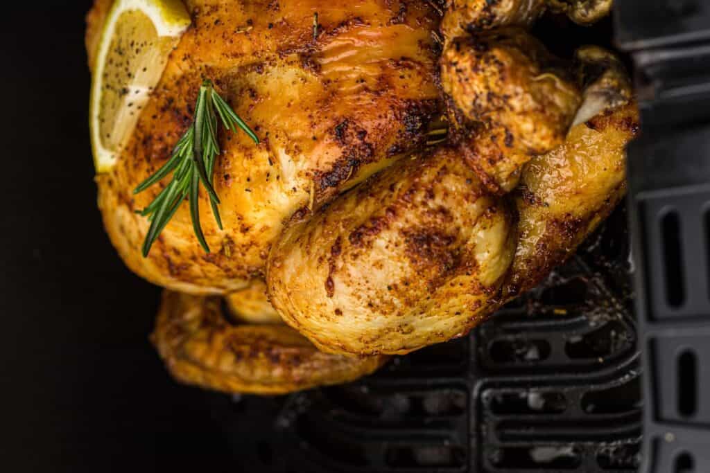 Close-up of a cooked, seasoned whole chicken garnished with a rosemary sprig and a lemon slice, resting in a kitchen appliance.