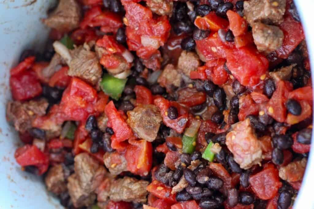 A close-up of a dish with cooked diced tomatoes, black beans, and meat mixed together.