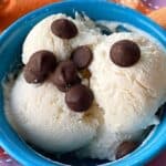 A blue bowl filled with three scoops of vanilla ice cream topped with a few chocolate chips, placed on a colorful, patterned cloth.
