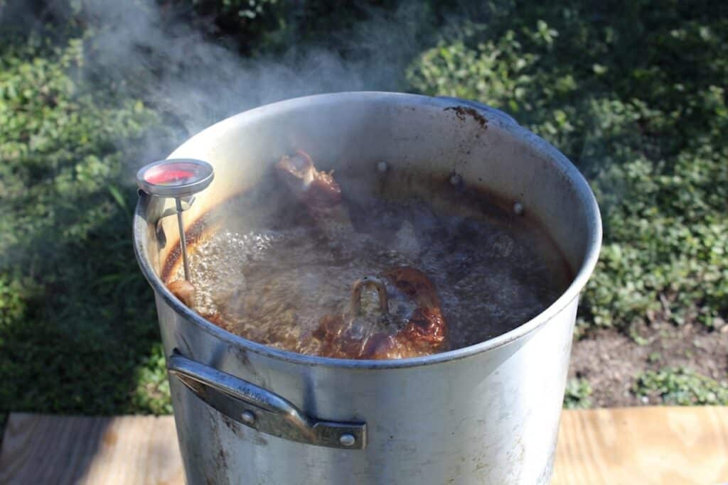 A large metal pot on an outdoor burner is filled with boiling oil and frying food. Steam rises, and a thermometer is clipped to the pot's side.