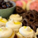 An assortment of snacks, including cupcakes with white frosting, chocolate-covered pretzels, strawberries, and a bowl of chocolate-covered nuts.
