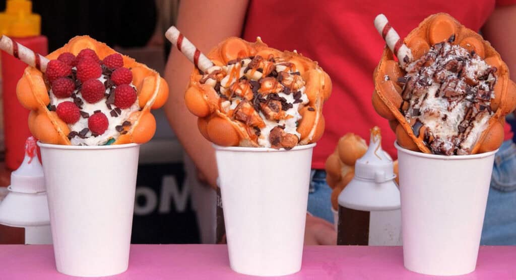 Three cups of bubble waffles, reminiscent of delightful state fair food, with assorted toppings including raspberries, chocolate pieces, and shredded coconut, each garnished with a chocolate stick wafer.