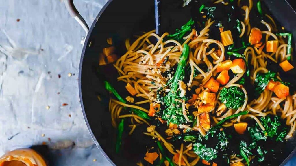 A pan of spaghetti mixed with kale, diced sweet potatoes, and garnished with sesame seeds.