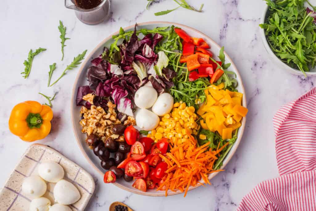 A colorful salad with various ingredients including mozzarella balls, chopped tomatoes, red bell peppers, corn, shredded carrots, black olives, and mixed greens, placed on a marble surface.