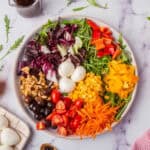 A colorful salad with various ingredients including mozzarella balls, chopped tomatoes, red bell peppers, corn, shredded carrots, black olives, and mixed greens, placed on a marble surface.