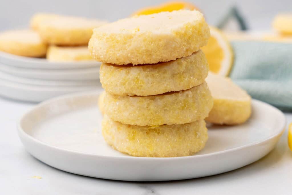 A stack of four round lemon cookies sits on a white plate, with more cookies and a lemon in the background.