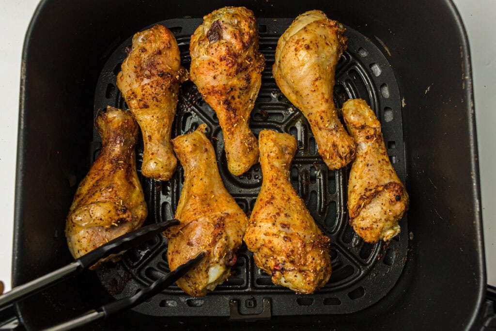 Six seasoned chicken drumsticks are arranged in an air fryer basket, with a pair of tongs holding one of them. The chicken has a cooked, browned appearance.