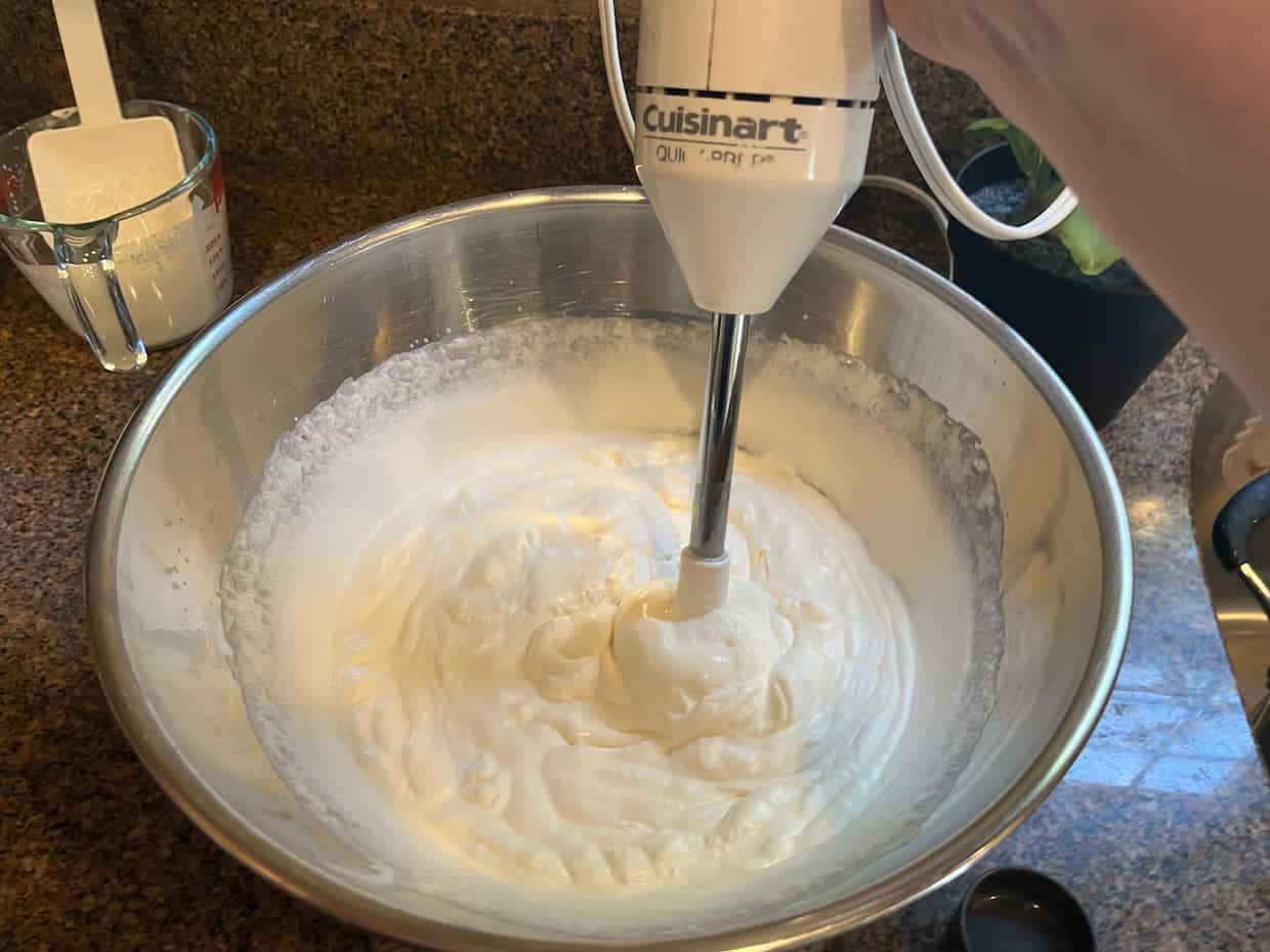 A hand using a Cuisinart immersion blender to whip cream in a large stainless steel bowl on a kitchen countertop. A measuring cup and a rubber spatula are in the background.