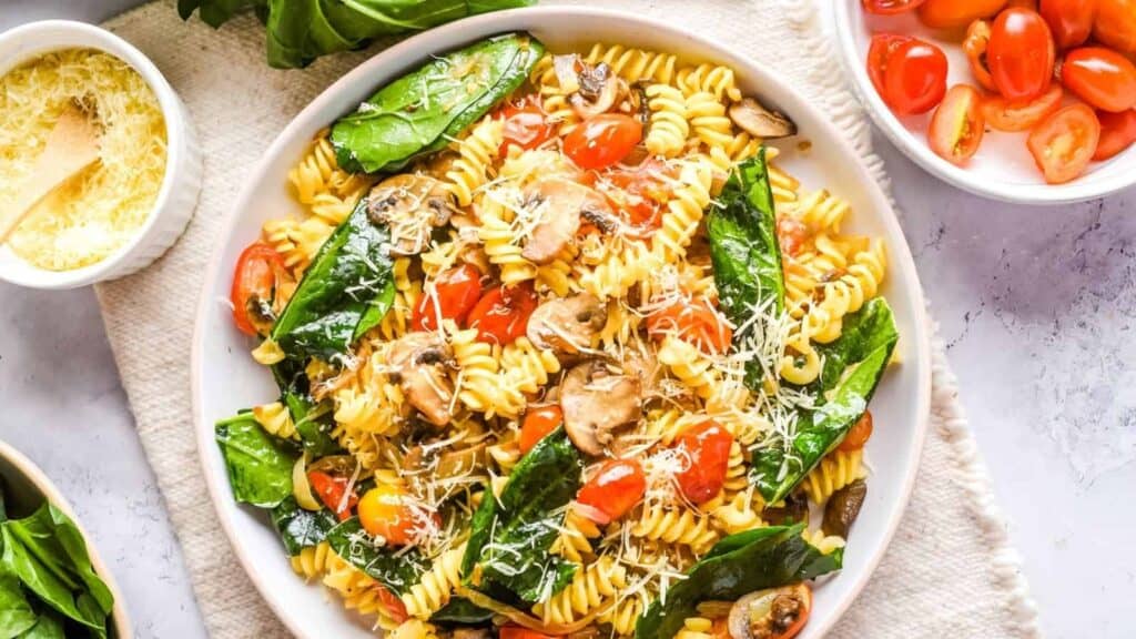 A plate of fusilli pasta with spinach, tomatoes, mushrooms, and grated cheese, surrounded by bowls of cherry tomatoes, spinach, and grated cheese.