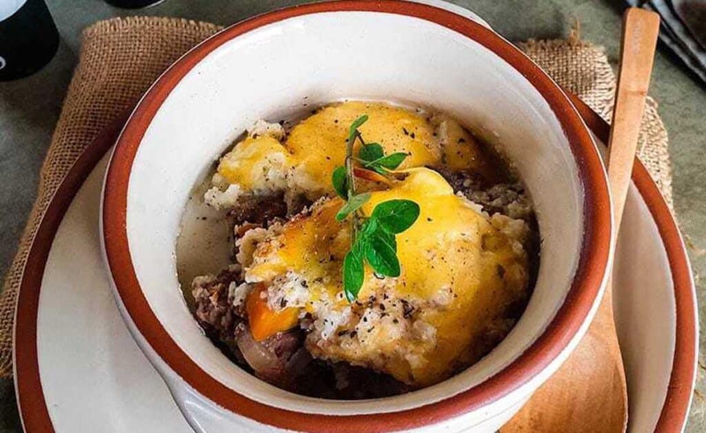 A ceramic bowl filled with a baked dish topped with melted cheese and garnished with green herbs. A wooden spoon rests beside it on a burlap cloth.