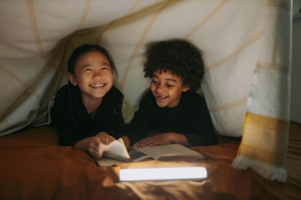 Two children smiling under a blanket fort with a book and a flashlight, creating a cozy and playful atmosphere.