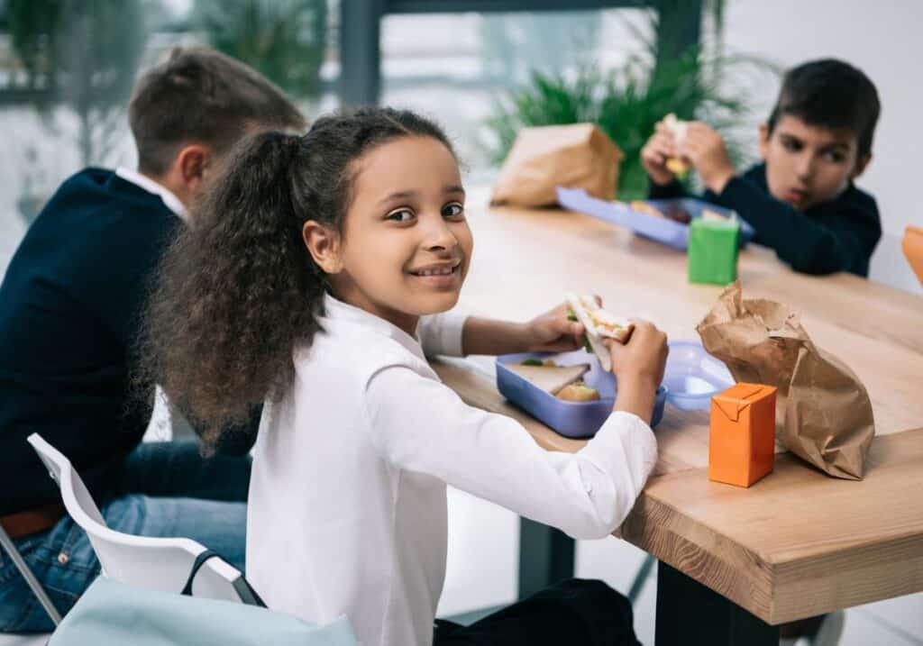 Three children are sitting at a table eating lunch. The girl in the foreground is looking at the camera and smiling while holding a sandwich, demonstrating one of her favorite lunch box hacks.