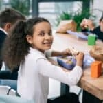 Three children are sitting at a table eating lunch. The girl in the foreground is looking at the camera and smiling while holding a sandwich, demonstrating one of her favorite lunch box hacks.