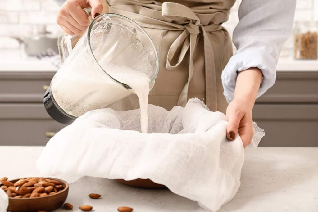 A person in an apron pours blended almond mixture through a cheesecloth into a bowl to make almond milk. A bowl of whole almonds is on the counter.