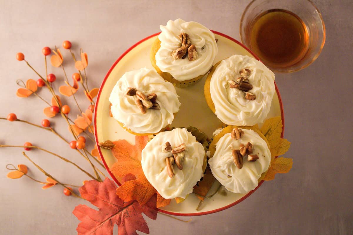 A plate of five cupcakes with cream frosting, decorated with nuts, is placed next to a sprig of orange berries and autumn leaves on a light gray surface. A small glass cup of tea is also nearby.
