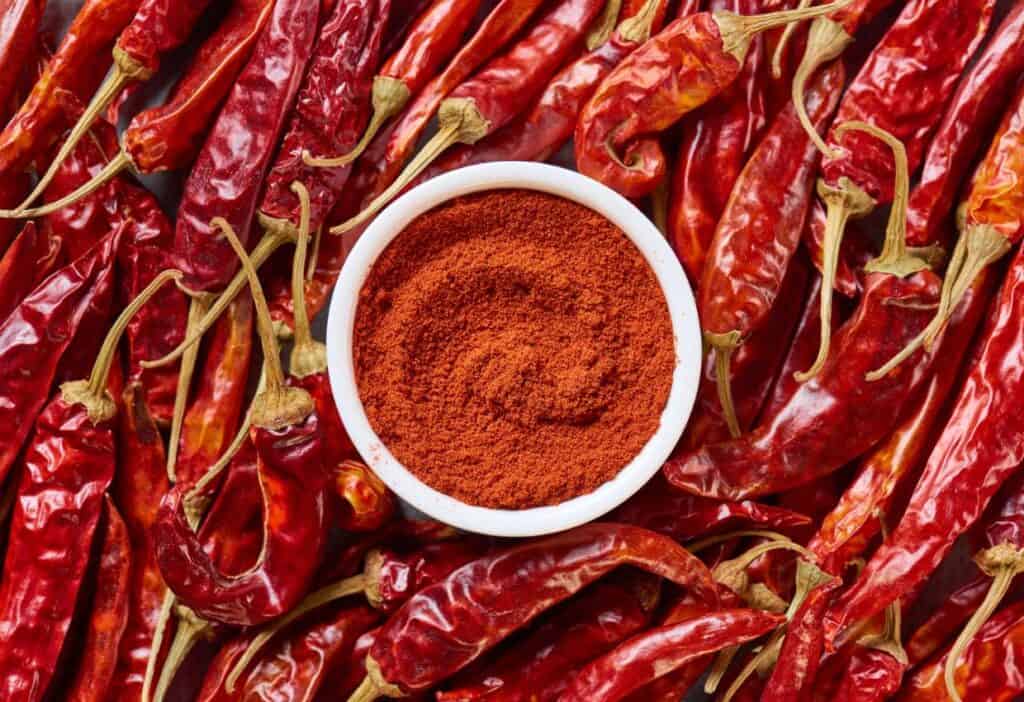 A white bowl filled with ground red chili powder is placed on a bed of whole dried red chilies.