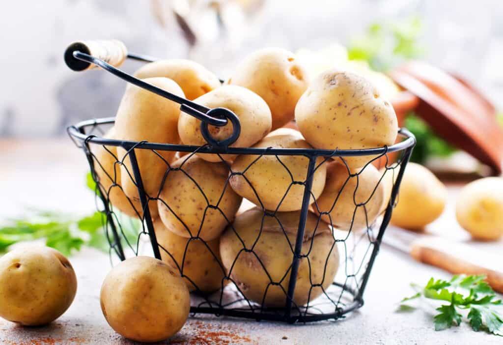 A wire basket filled with unpeeled potatoes on a counter, with some parsley and a knife nearby.