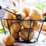 A wire basket filled with unpeeled potatoes on a counter, with some parsley and a knife nearby.