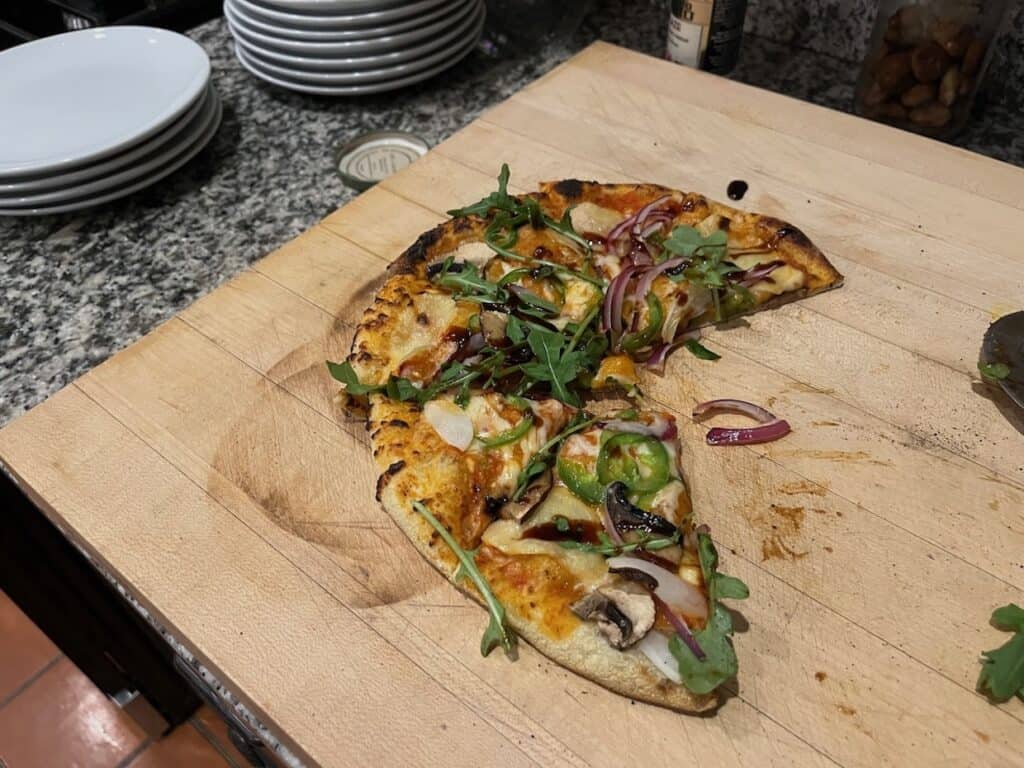 A partially eaten pizza topped with arugula, red onion, mushrooms, and jalapeños on a cutting board next to a stack of white plates.