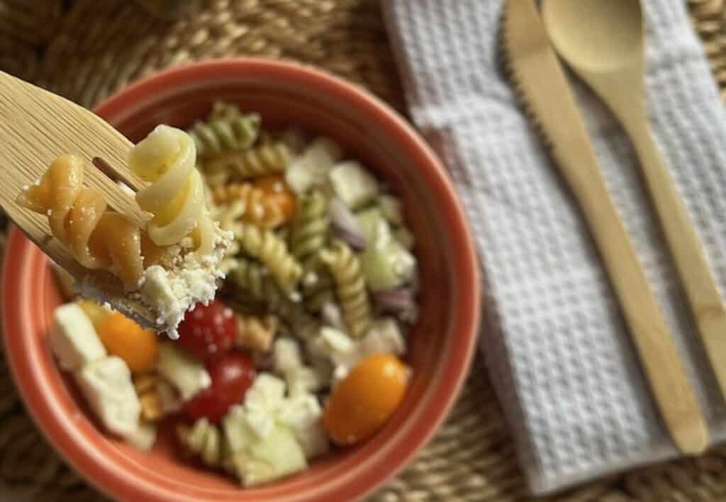 A fork with pasta near a bowl of mixed pasta salad containing tomatoes, feta cheese, and other vegetables. A wooden spoon and fork rest on a white cloth nearby.