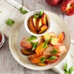 Plate with cooked chicken pieces garnished with fresh peach slices and herbs, served with a small bowl of peach slices and a jar of sauce on a wooden table, accompanied by two whole peaches.