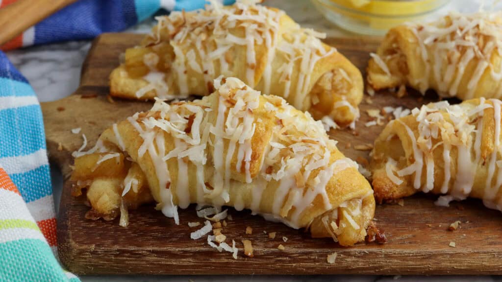 Four coconut-glazed pastry croissants on a wooden board, garnished with shredded coconut, and accompanied by a colorful striped cloth.