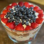 A layered trifle dessert in a glass bowl, topped with whole strawberries and blueberries. The layers include cake, cream, and fruit.