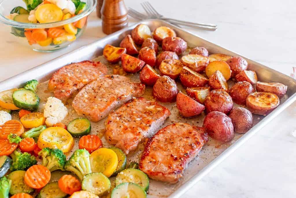 A sheet pan with cooked pork chops, roasted red potatoes, and an assortment of grilled vegetables including broccoli, cauliflower, carrots, and zucchini. A bowl of additional vegetables is in the background.
