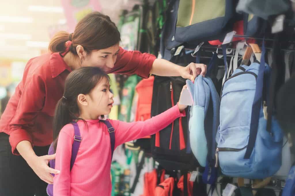 A woman and a young girl are in a store looking at a display of backpacks. The woman is pointing at a light blue backpack while the girl in a pink shirt looks on.