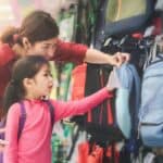 A woman and a young girl are in a store looking at a display of backpacks. The woman is pointing at a light blue backpack while the girl in a pink shirt looks on.