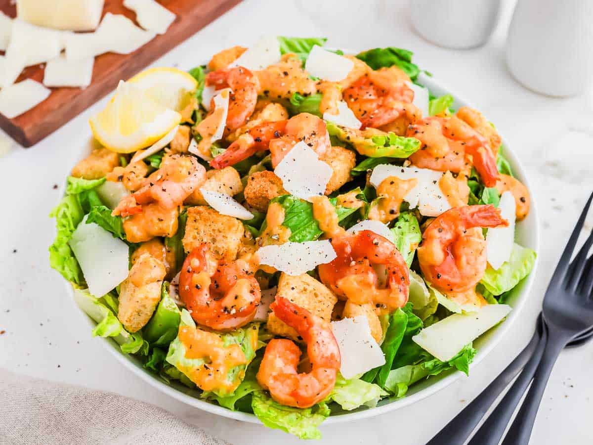 A fresh shrimp Caesar salad with romaine lettuce, shrimp, croutons, shaved parmesan cheese, and Caesar dressing is served in a white bowl with black utensils on a marble table.