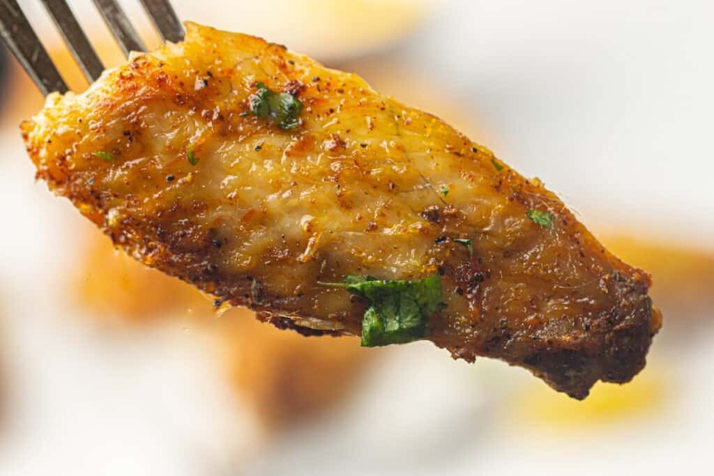 Close-up of a seasoned and cooked chicken wing held by a fork, garnished with small green herbs.