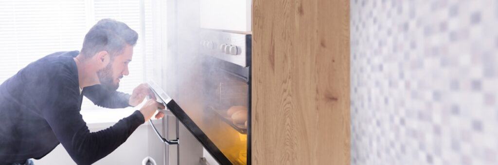 A man looks into a smoky oven in a kitchen, checking the contents while holding the oven door slightly open.