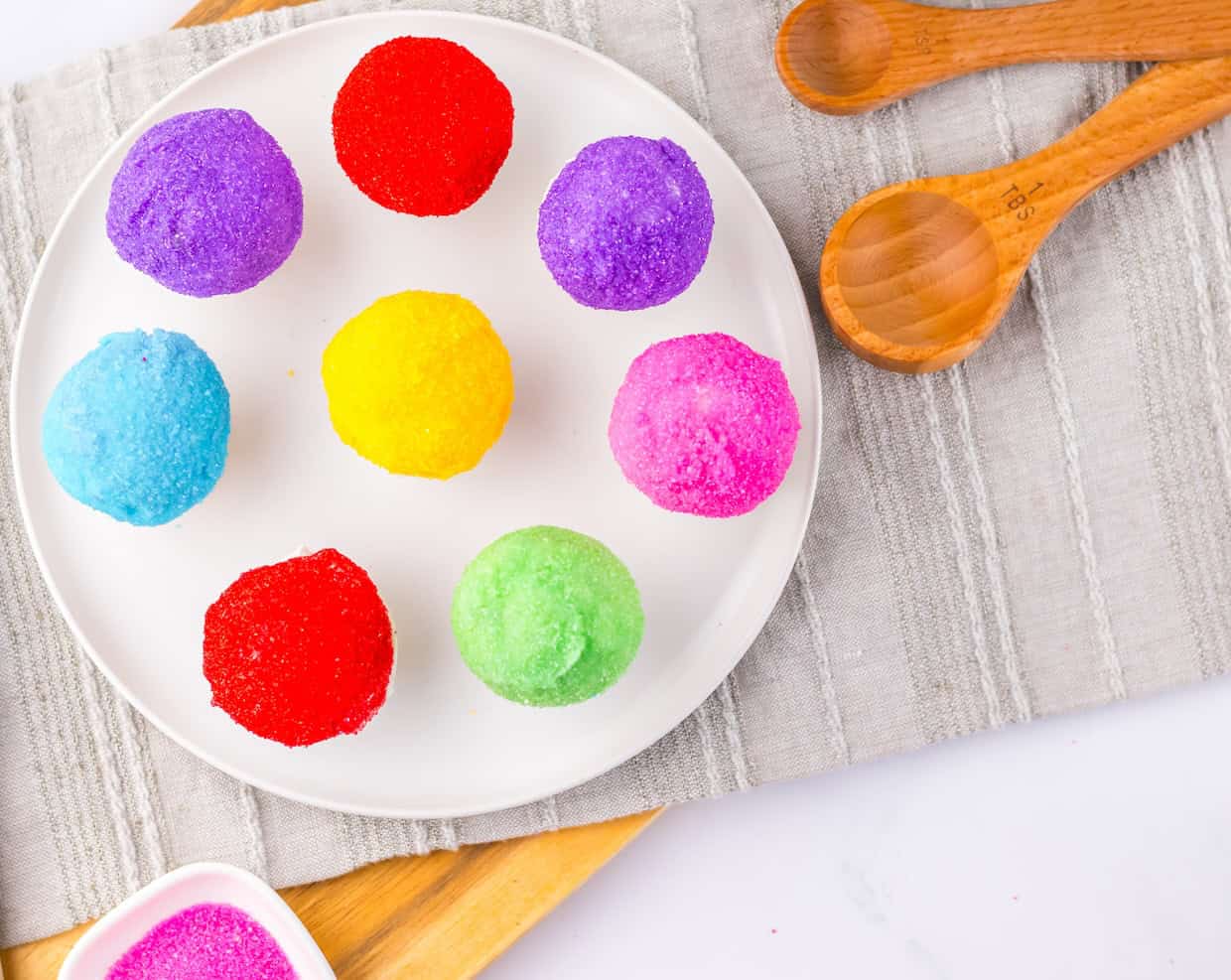 A white plate with nine round, colorful balls of sugar-coated dough on a gray striped cloth; two wooden scoops lie nearby.