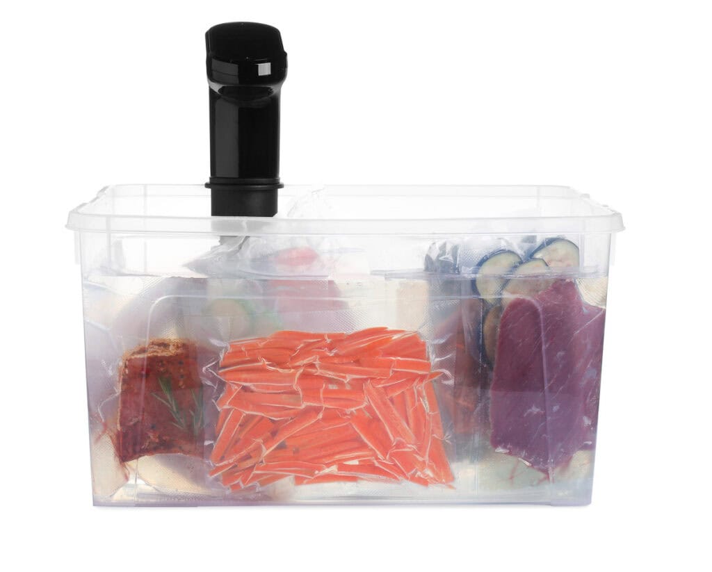 A sous vide cooker with multiple vacuum-sealed bags of sliced carrots, meat, and vegetables submerged in water inside a transparent container.