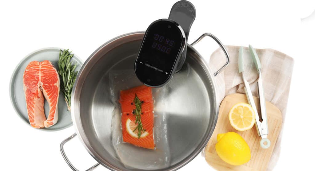 A sous vide machine cooking a vacuum-sealed salmon fillet with herbs and lemon in a pot. Nearby, a plate with raw salmon, rosemary, a cutting board with a lemon, and kitchen tongs are visible.