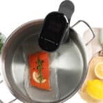 A sous vide machine cooking a vacuum-sealed salmon fillet with herbs and lemon in a pot. Nearby, a plate with raw salmon, rosemary, a cutting board with a lemon, and kitchen tongs are visible.
