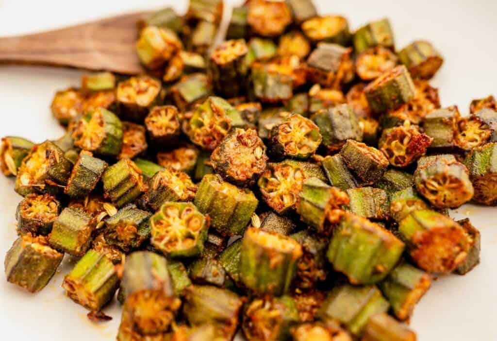 A plate of cooked, seasoned okra pieces, reminiscent of classic southern food, with a wooden spoon in the background. The okra appears to be roasted or sautéed, displaying a golden-brown color.
