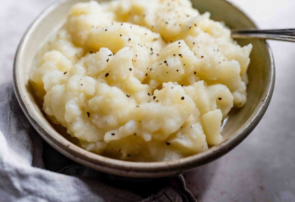 A close-up view of creamy mashed potatoes in a bowl, topped with freshly ground black pepper. A spoon is partially visible on the right side of the bowl, evoking the charm of southern food. A cloth napkin is on the left side.