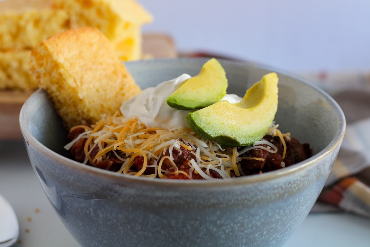 A bowl of chili topped with shredded cheese, sour cream, and avocado slices, accompanied by pieces of cornbread.