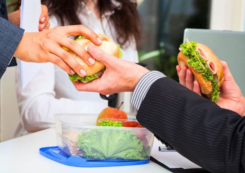 People exchanging sandwiches in an office setting; one person also has a container with salad and fruit on the table.