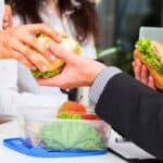 People exchanging sandwiches in an office setting; one person also has a container with salad and fruit on the table.