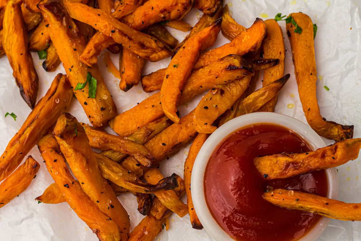 A plate of cooked sweet potato fries with black pepper seasoning is arranged on parchment paper beside a small container of ketchup.
